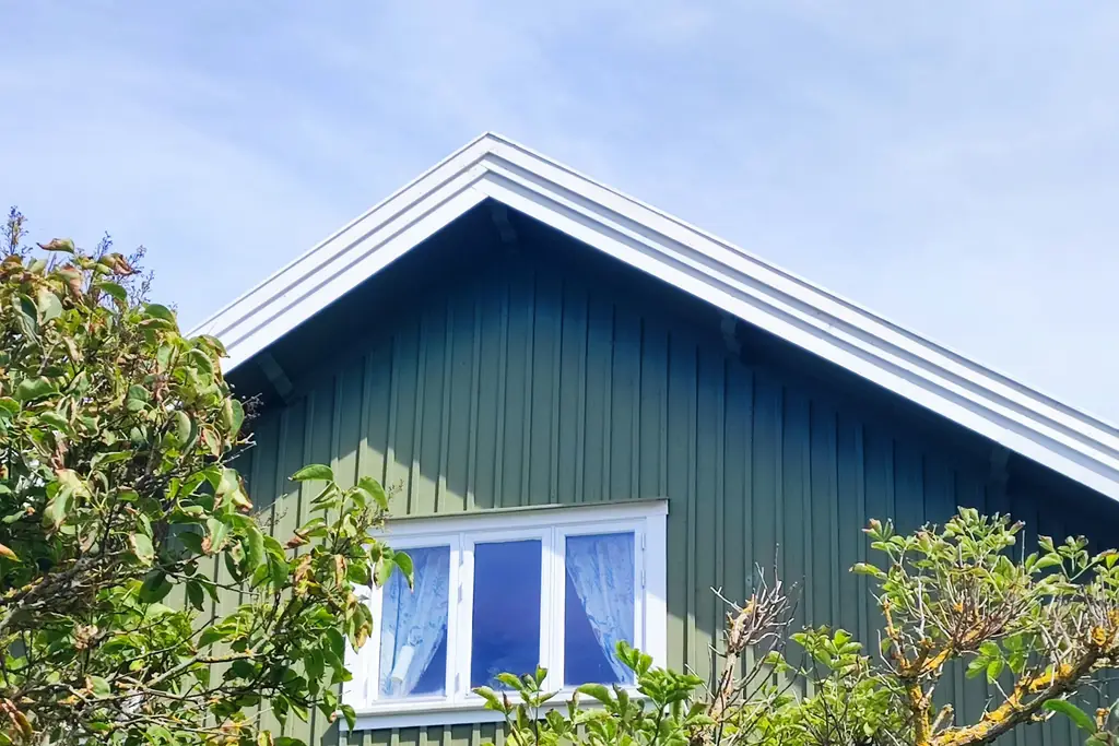 Grönt trähus med vita fönster bakom träd och buskar
